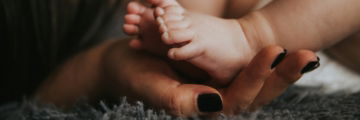infant-s-feet-being-held-by-a-woman-s-hand-with-painted-and-manicured-hands-resting-on-a-gray-blanket.jpg Infant's feet being held by a woman's hand with painted and manicured hands resting on a gray blanket